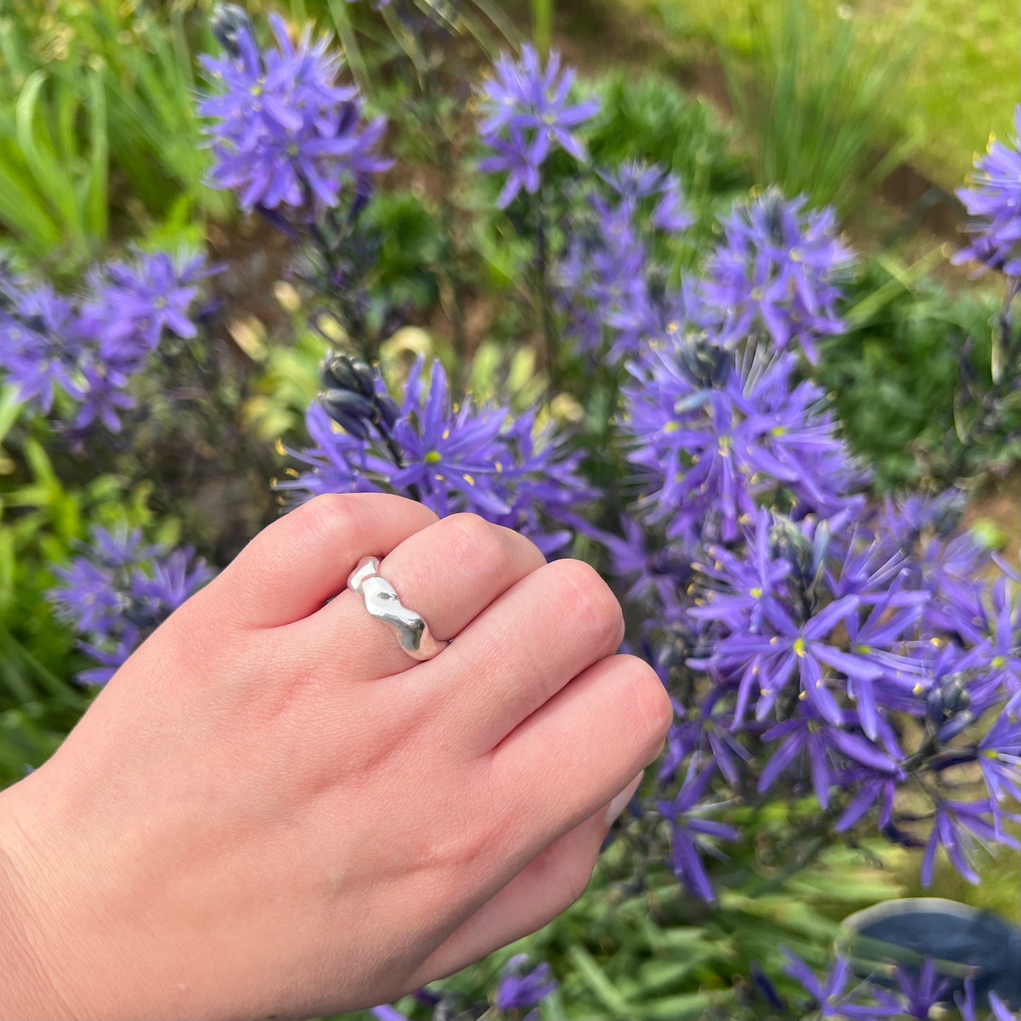 The wave ring in shiny sterling silver on a hand with a background of purple flowers.