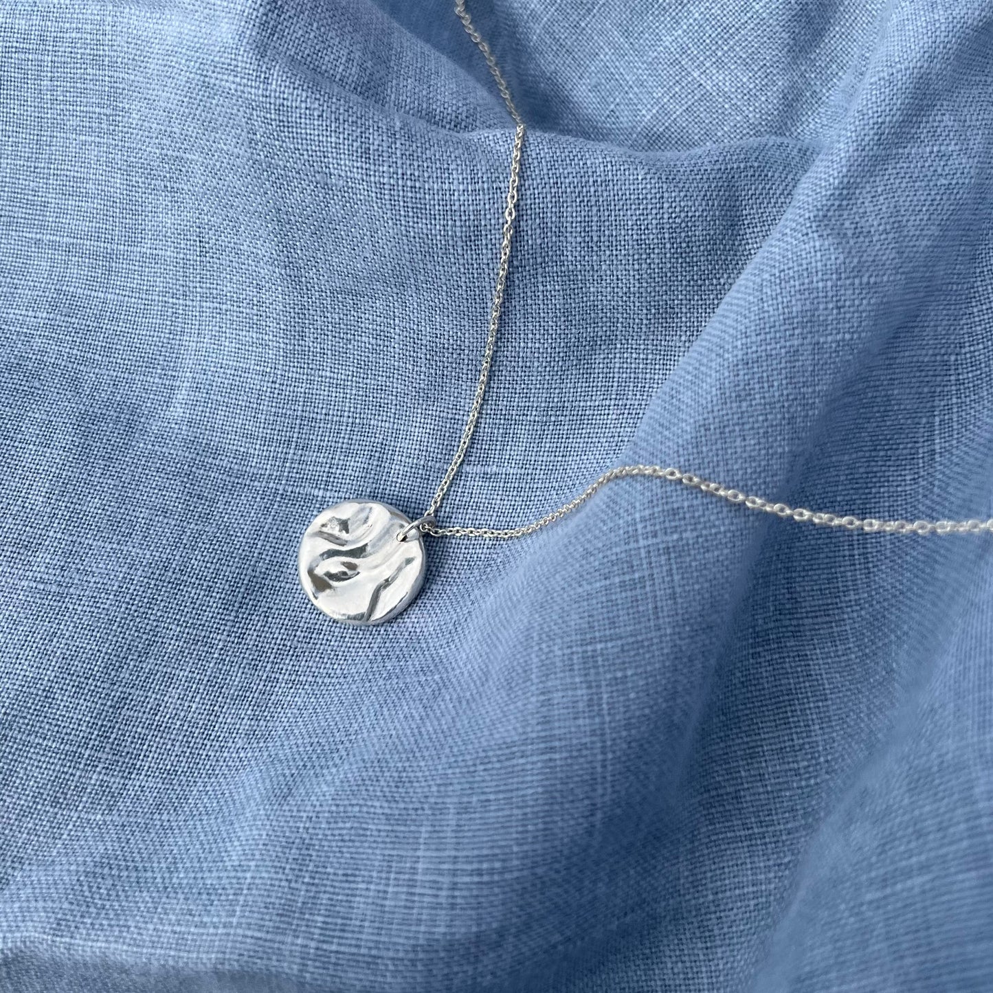 A shiny sterling silver 'Rock-pool pendant". The pendant is on a belcher chain and the background is of blue linen.