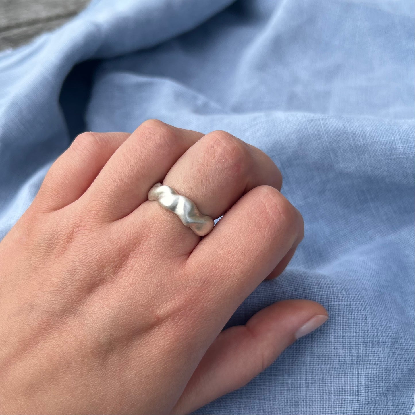 The wave ring in matt sterling silver on a hand with a background of blue linen.