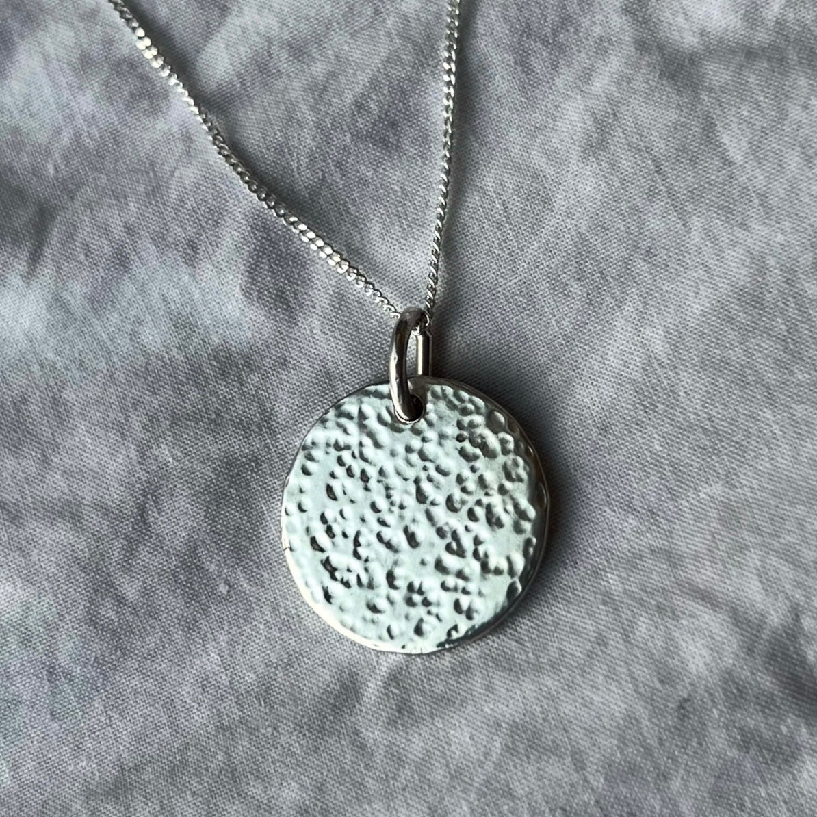 A sterling silver hammered texture charm on a chain on a white linen background.