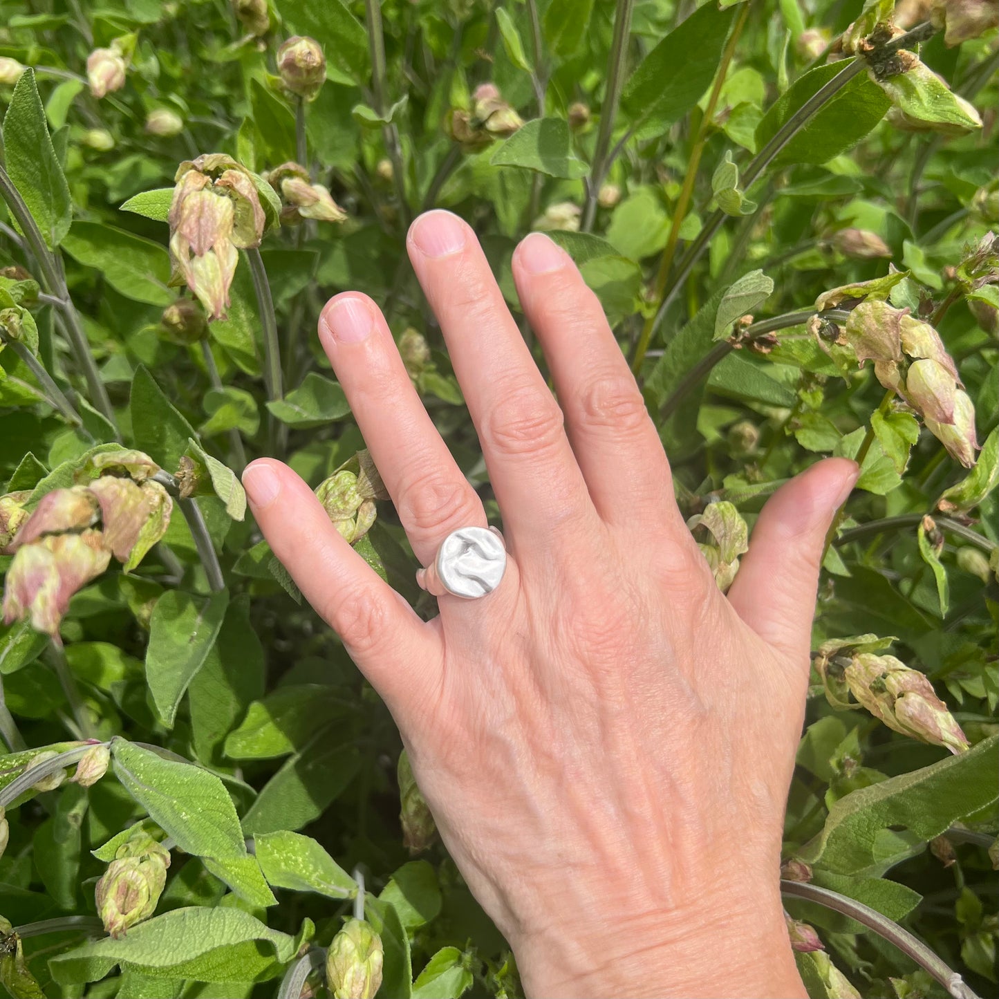 A matt sterling silver splash ring on a hand with a shrub in the background.
