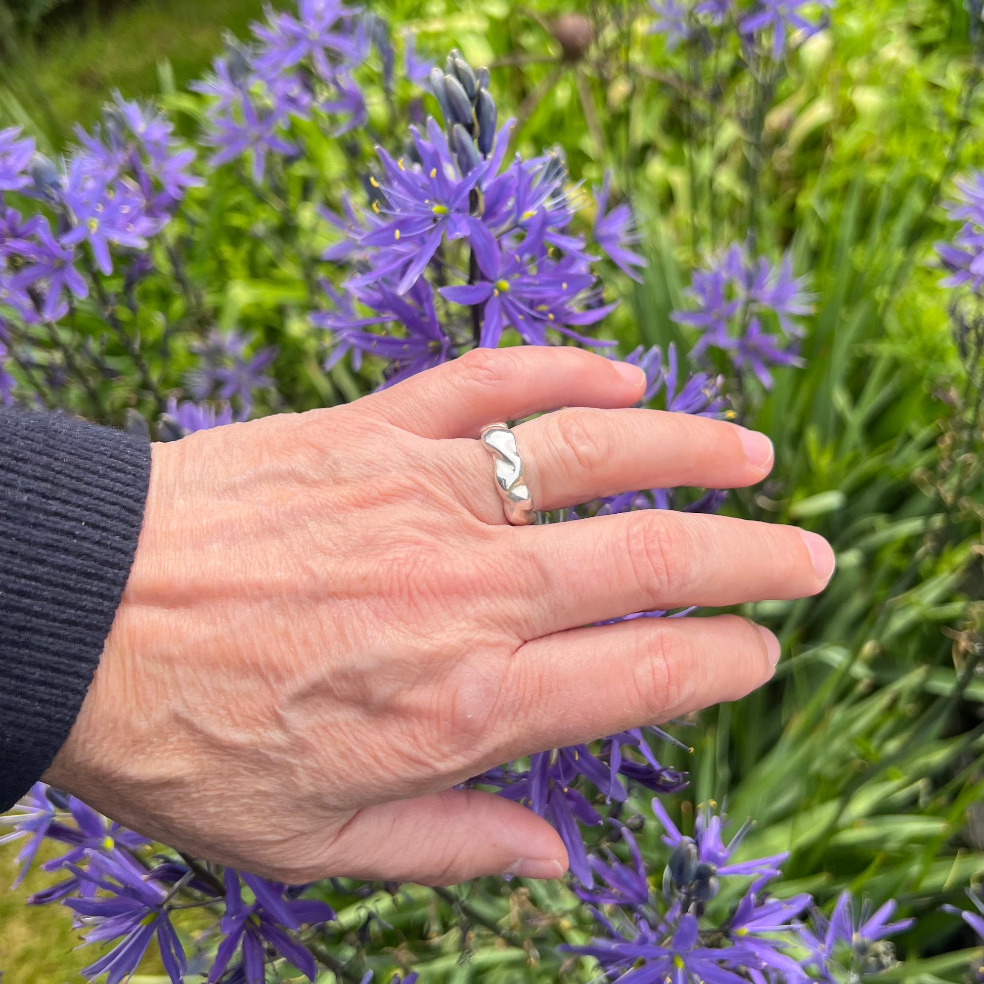 The wave ring in shiny sterling silver on a hand with a background of purple flowers.