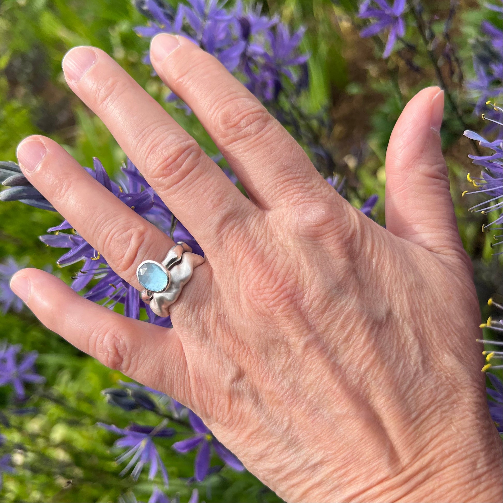 A matt wave ring and a topaz set bubble ring on a hand with a background of purple flowers.