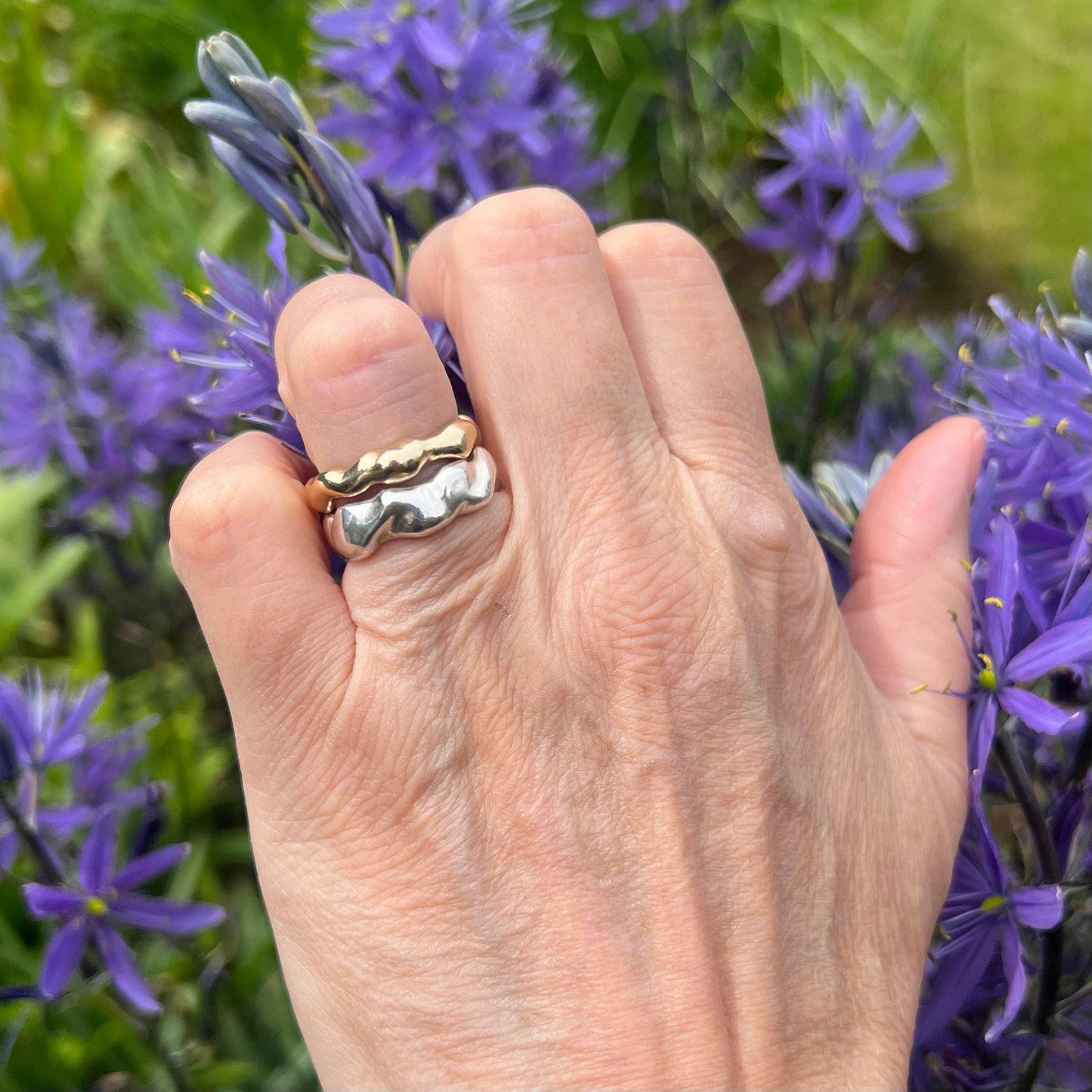 A sterling silver wave band and a gold bubble ring on a hand with a background of purple flowers.