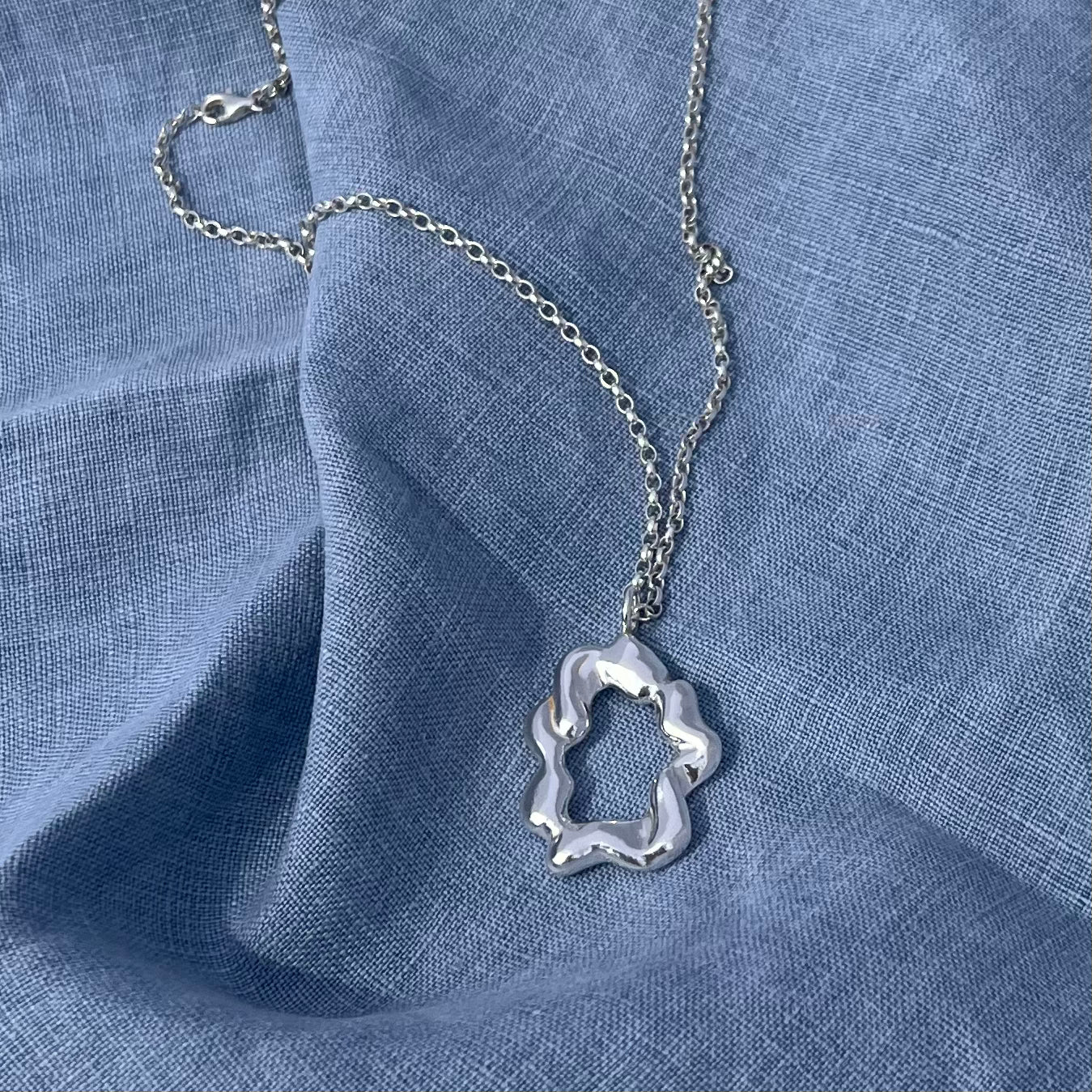 The 'Cloud pendant'. A sterling silver abstract shaped pendant on a silver belcher chain. The background is blue linen.