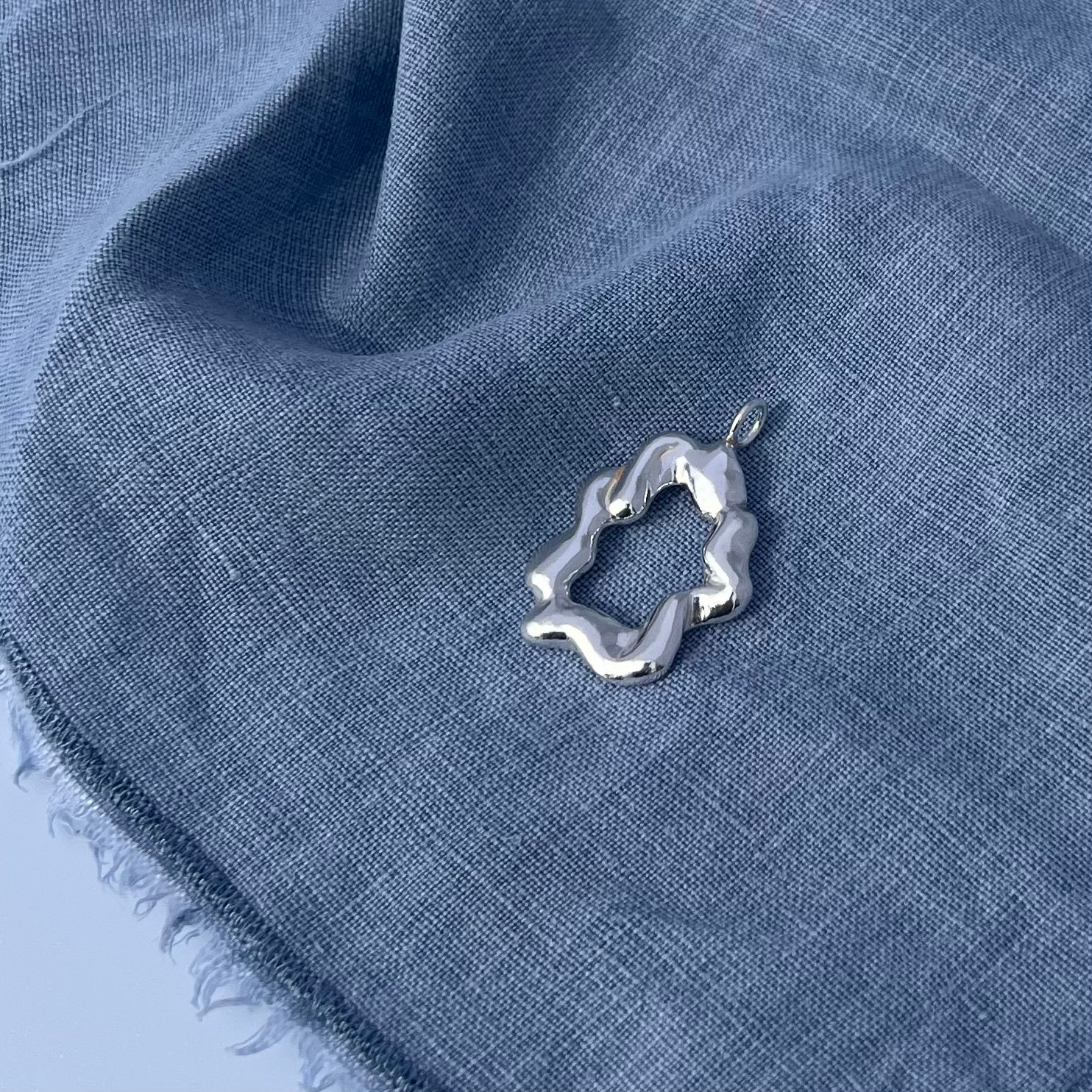 The 'Cloud pendant'. A handmade, sterling silver abstract shaped pendant. The background is blue linen.