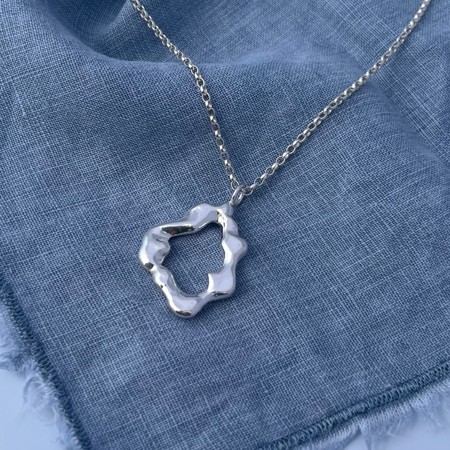 The 'Mist pendant'. A sterling silver abstract shaped pendant on a silver belcher chain. The background is blue linen.