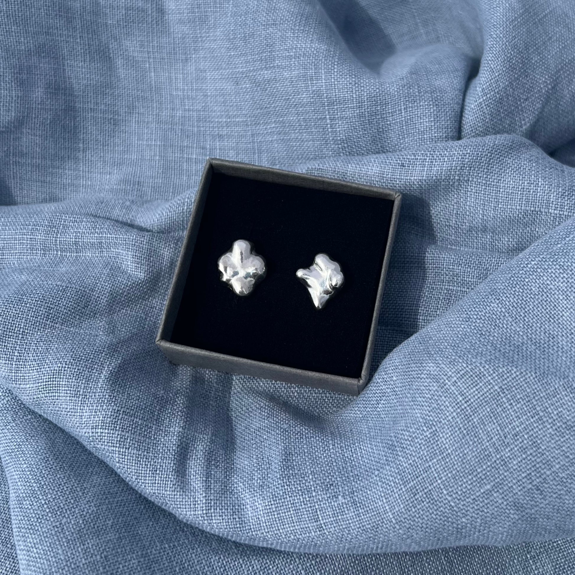 The sterling silver swirling stud earrings in a black box on a blue linen background.
