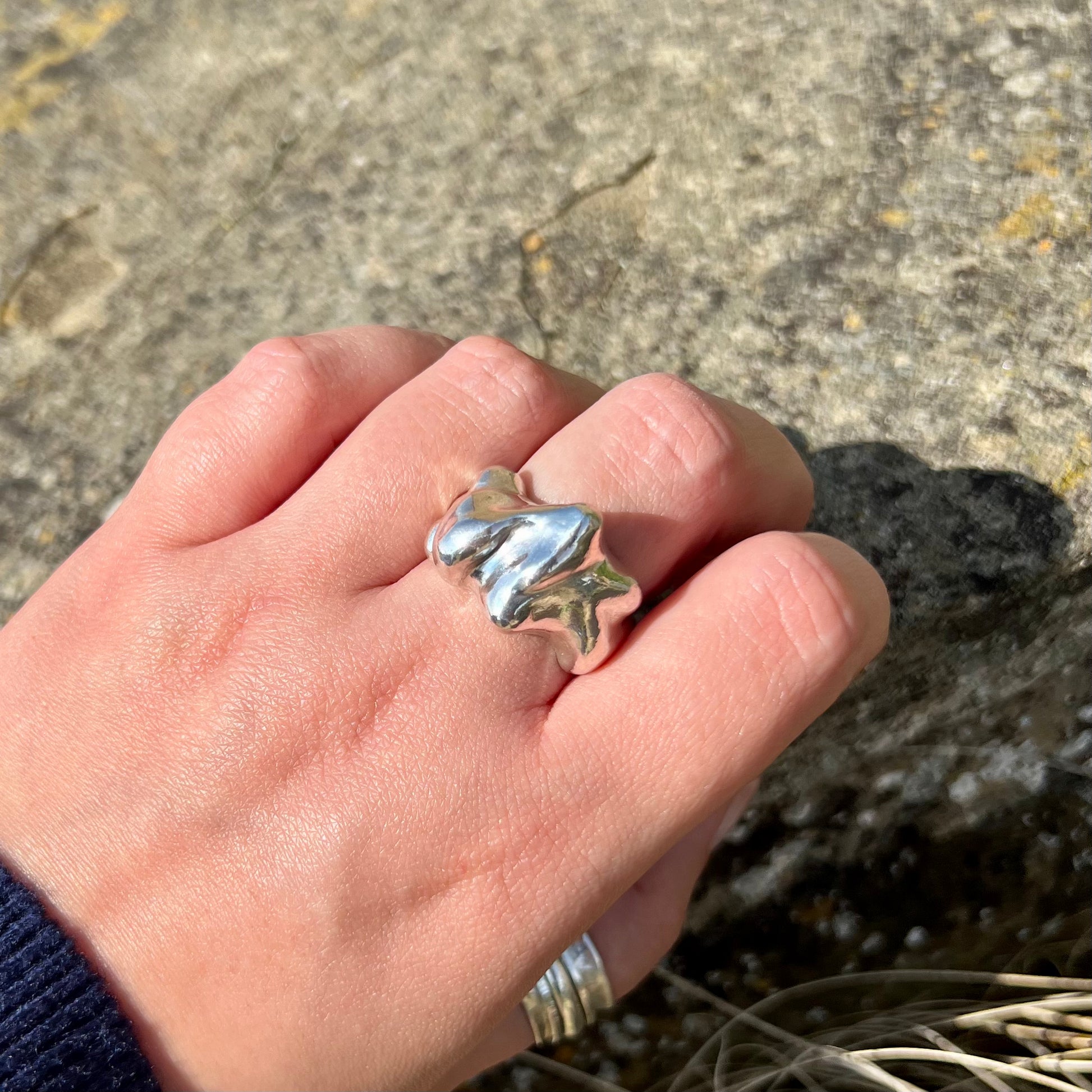 The Craggy ring. A chunky statement ring handmade in sterling silver which is photographed on a hand. The background is of a rock.