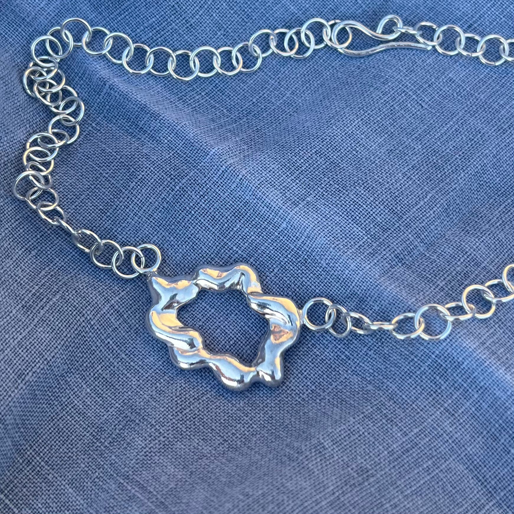 A zoomed in photograph of a chunky, statement handmade sterling silver bracelet on a blue linen background. The centrepiece of the bracelet is shaped like a cloud and it has a handmade hook clasp.