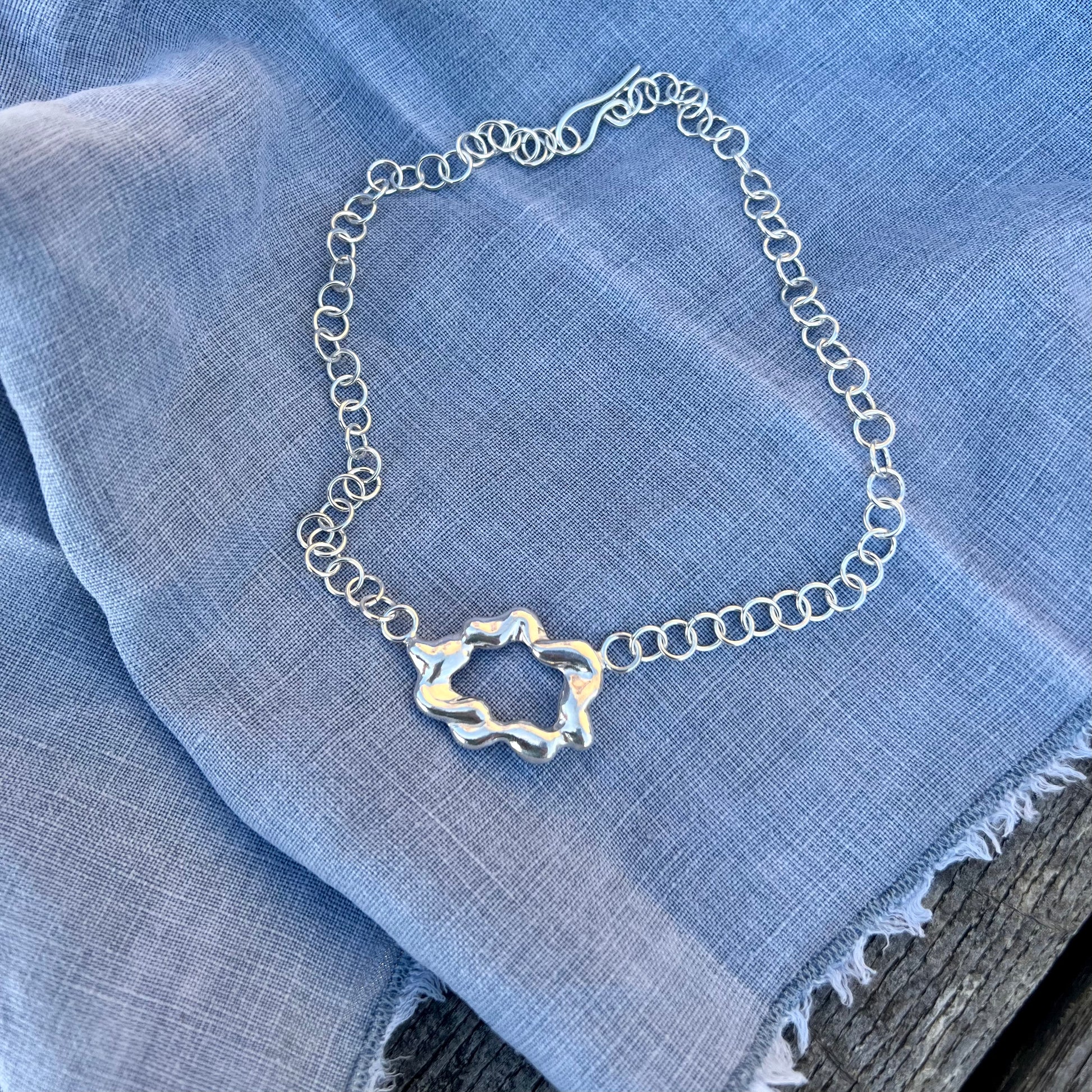 A chunky, statement handmade sterling silver necklace on a blue linen background. The centrepiece of the necklace is shaped like a cloud and it has a handmade hook clasp.