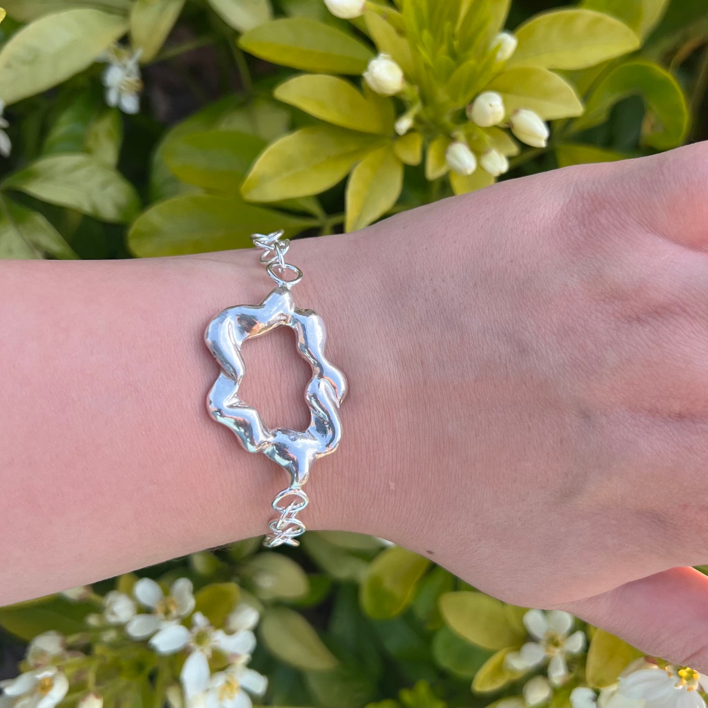 A chunky sterling silver bracelet on a wrist. The centrepiece of the bracelet is shaped like a cloud and the background is of a green shrub with white flowers.