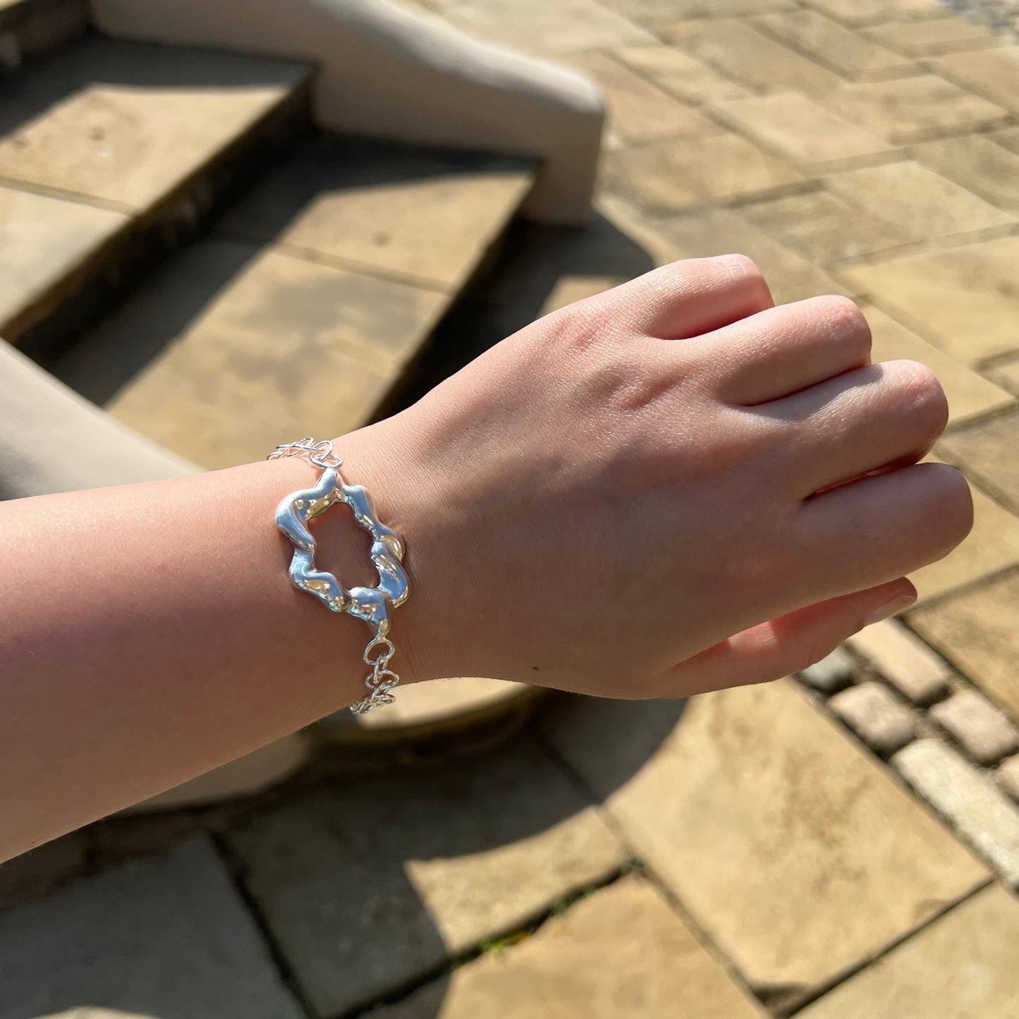 A handmade chunky sterling silver bracelet on a wrist. The centrepiece of the bracelet is shaped like a cloud and the background is of some stone steps and a patio.