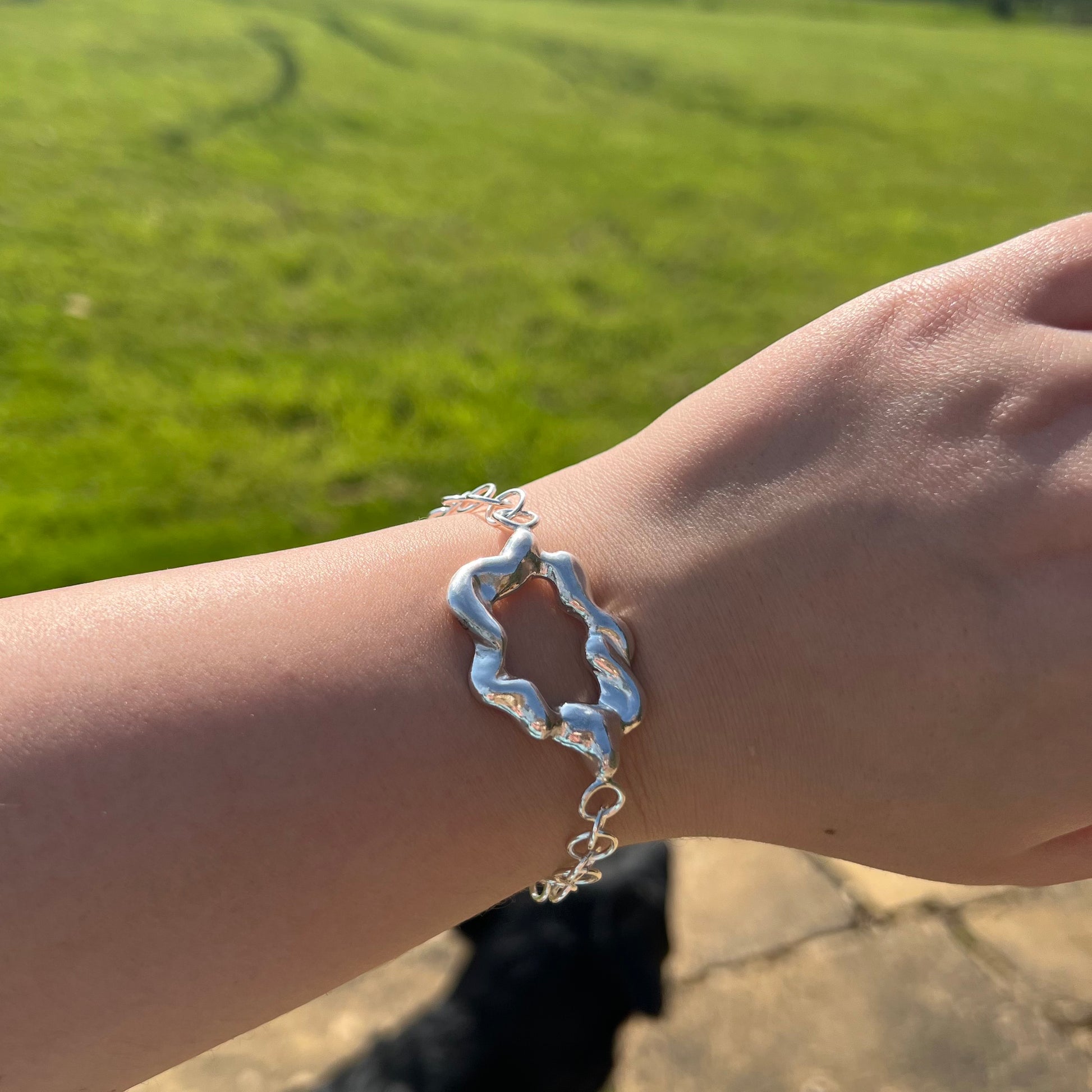 A chunky handmade sterling silver bracelet on a wrist. The centrepiece of the bracelet is shaped like a cloud and the background is of grass. a stone patio and a labrador.