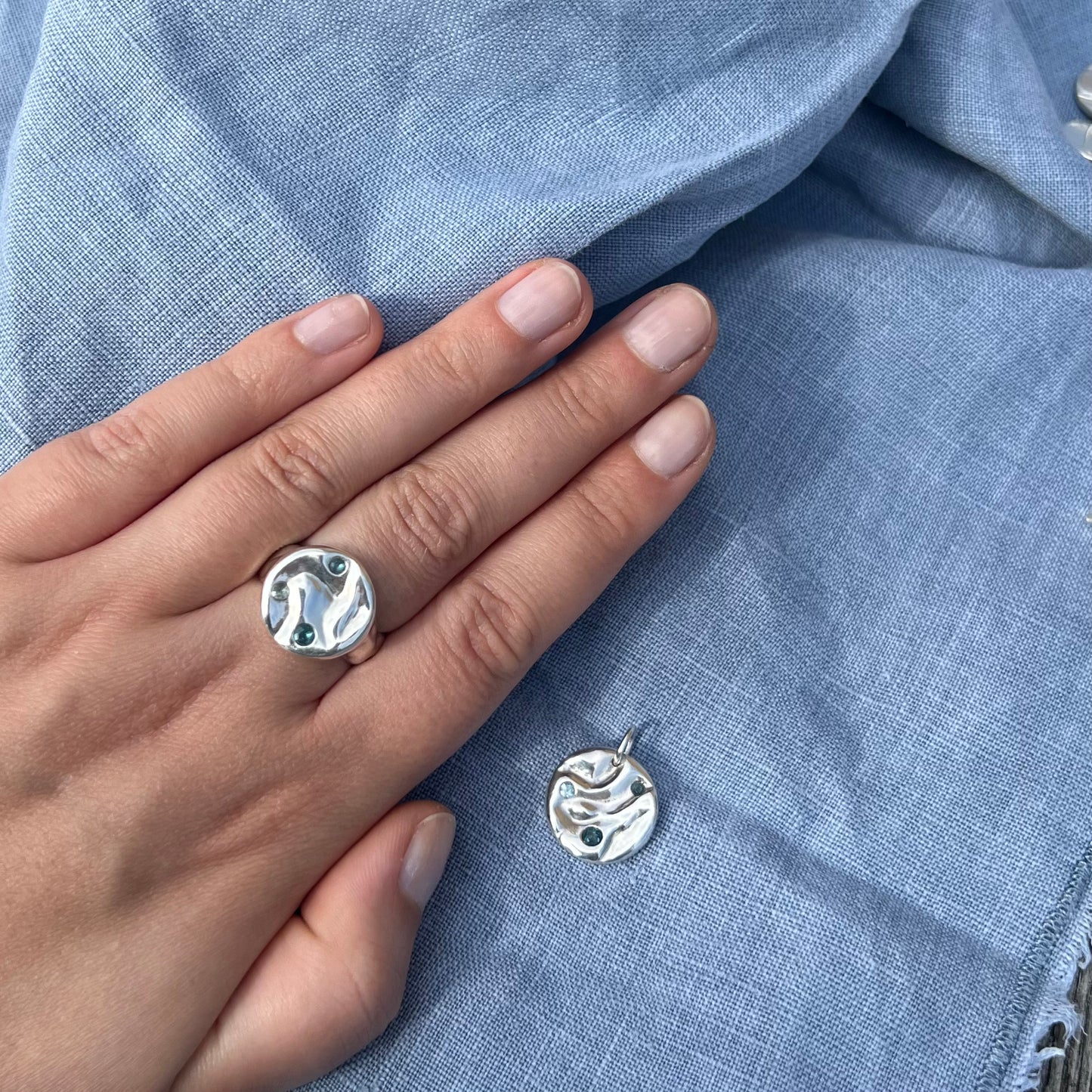 The triple topaz splash ring on a hand pictured next to the triple topaz rock pool pendant on a blue linen background.