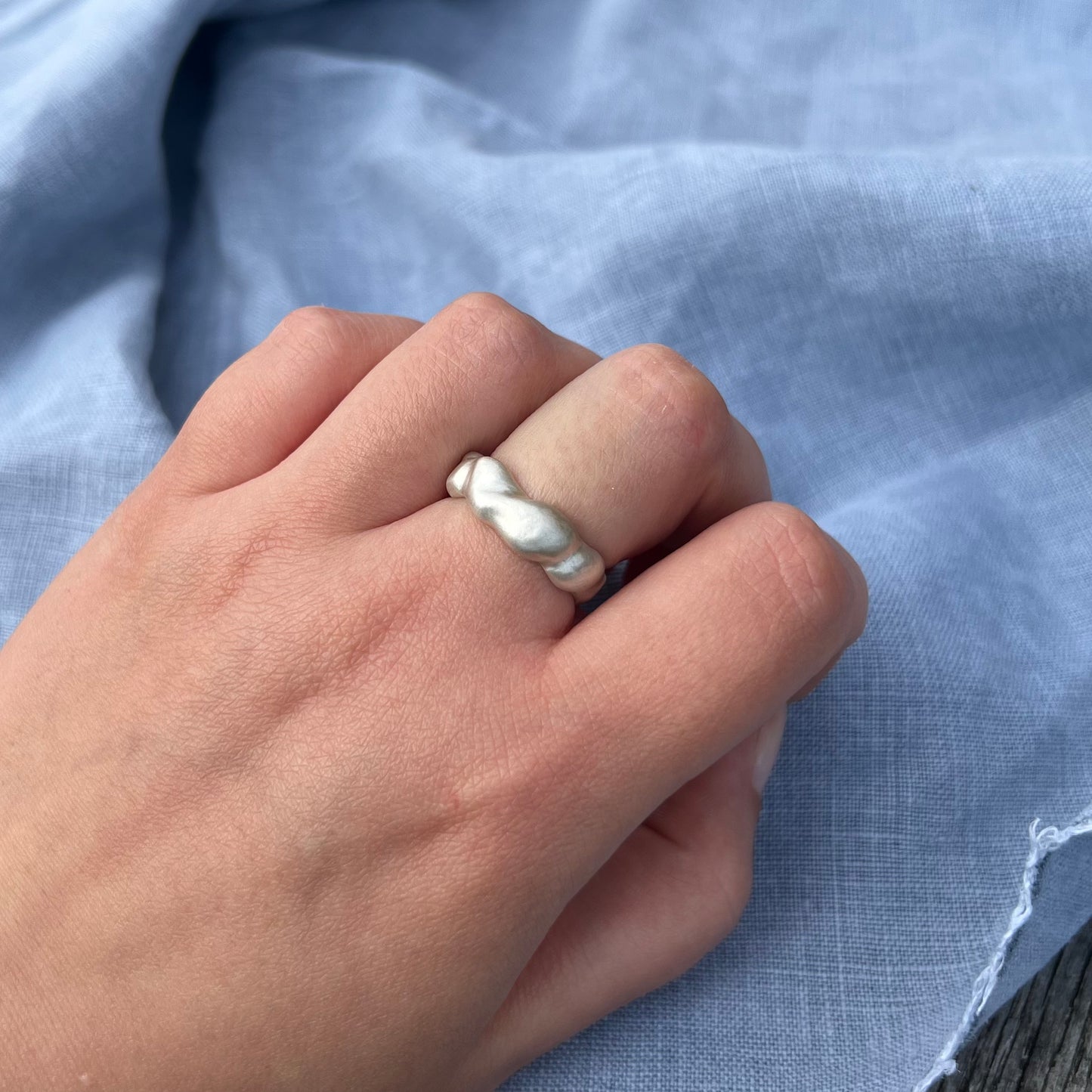 The wave ring in matt sterling silver on a hand with a background of blue linen.