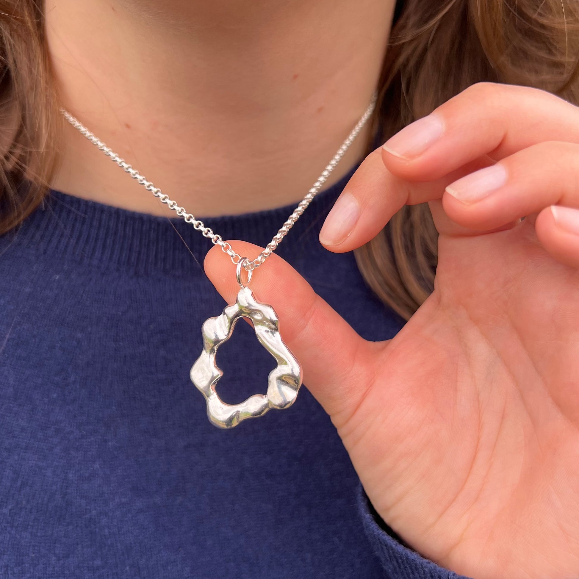 The 'Mist pendant'. A handmade, sterling silver abstract shaped pendant on a belcher chain. The necklace is pictured on a woman with a blue jumper on.