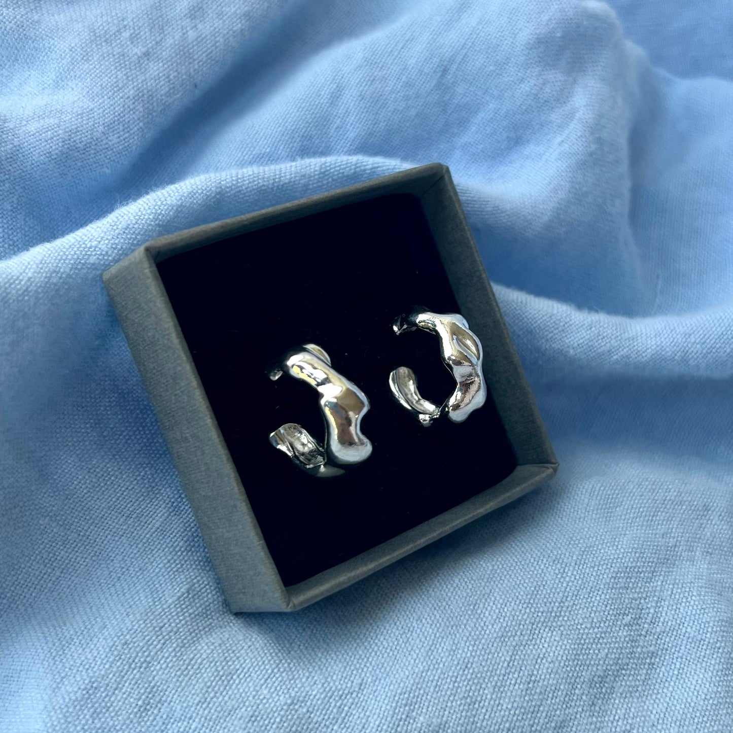 Small wavy silver earrings in a grey box on a blue background.