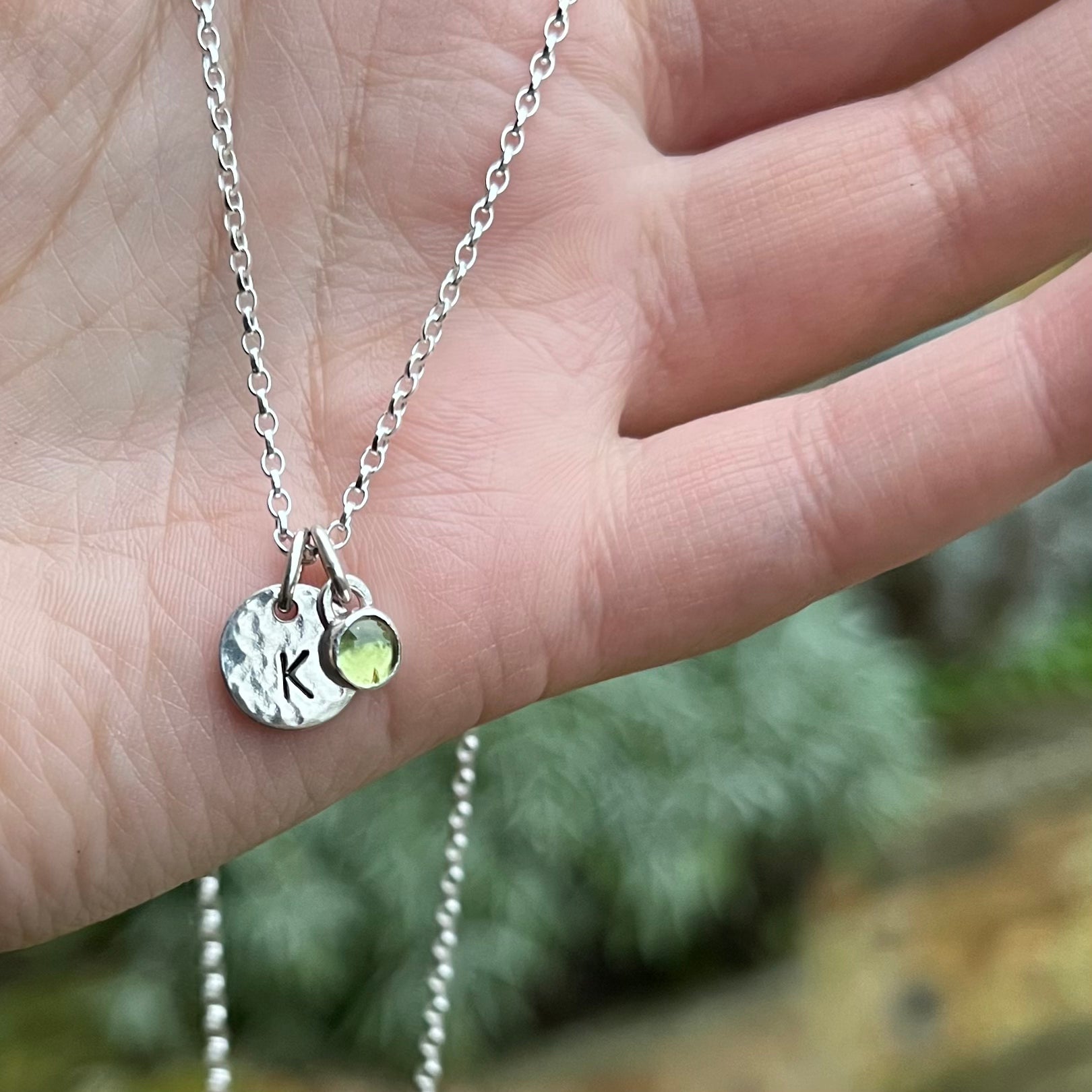A necklace with a sterling silver disc charm stamped with a K paired with a 5mm peridot stone charm.