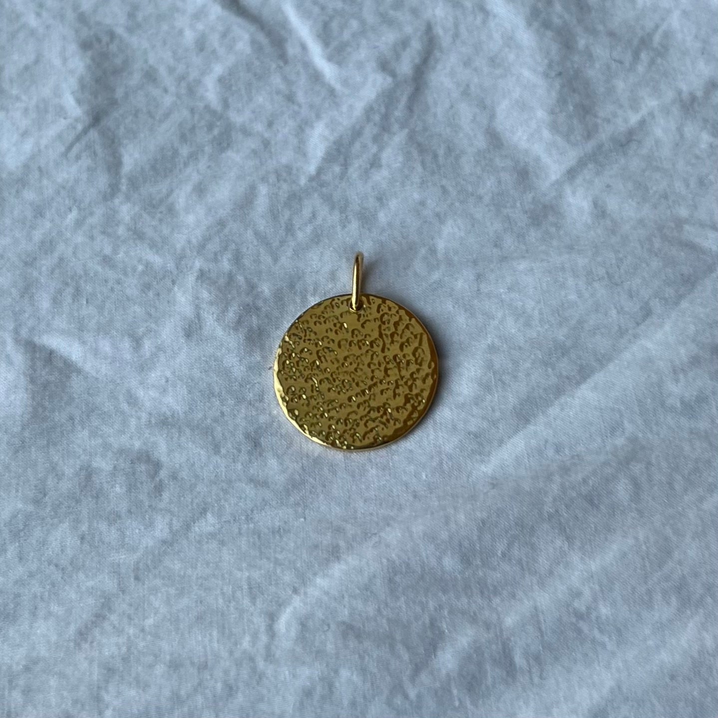 A gold vermeil hammered texture disc charm on a white linen background.