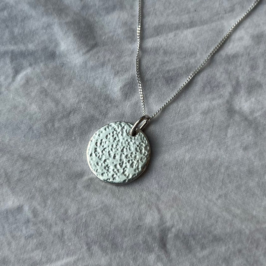 A sterling silver hammered texture charm on a chain on a white linen background.